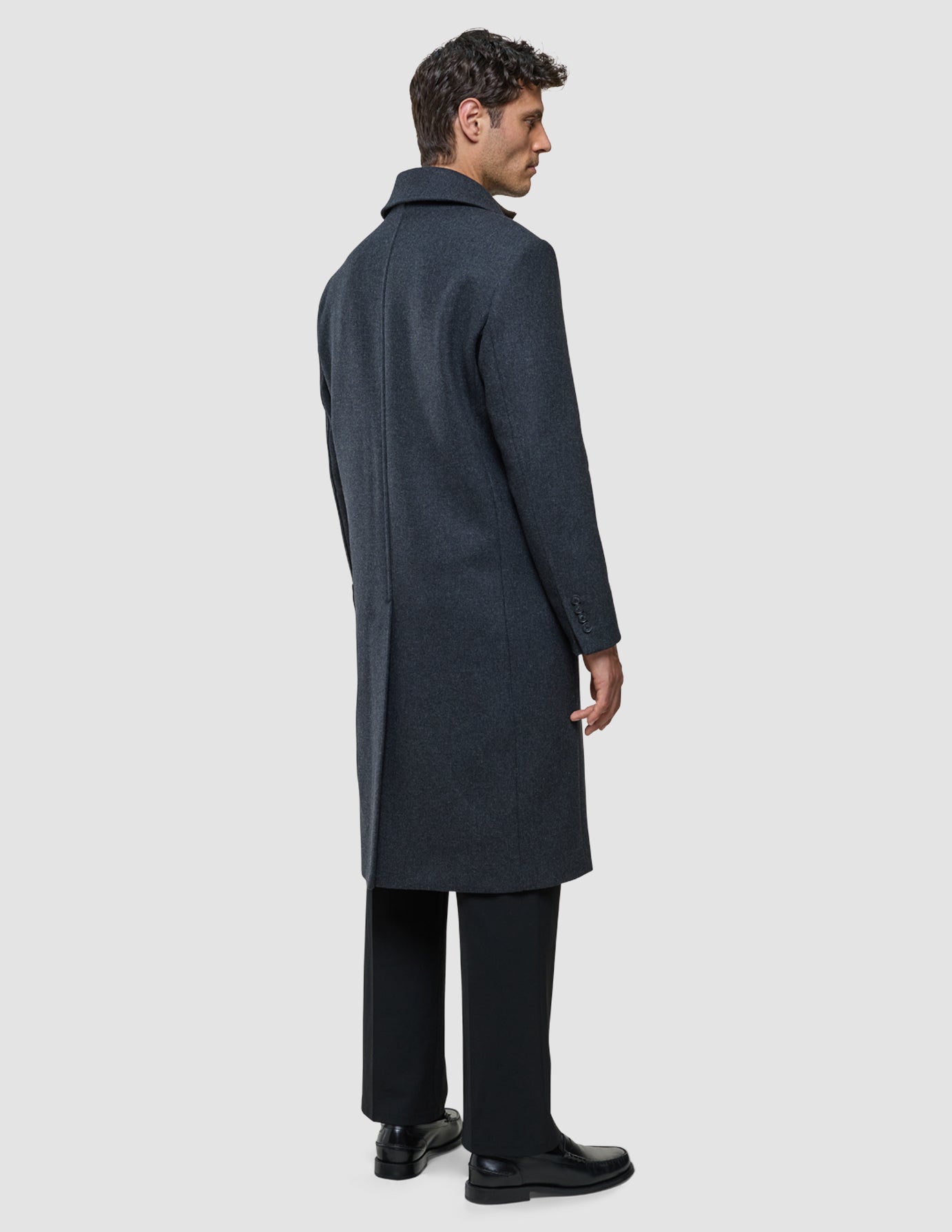 Ein Mann, von der Seite und von hinten gesehen, der einen dunkelgrauen Maxi-Wollmantel, eine schwarze Hose und schwarze Slipper trägt.